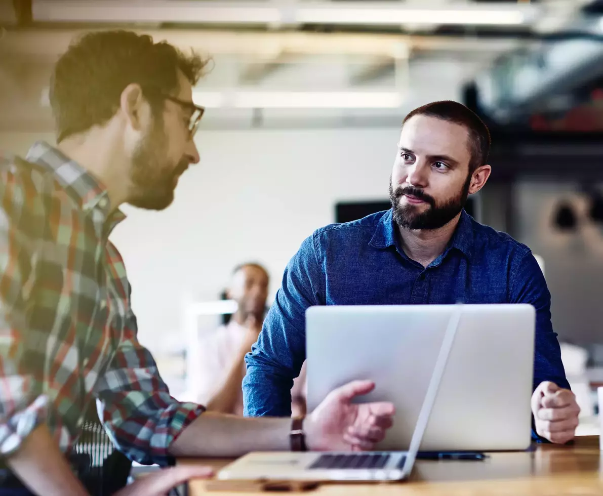 Two males talking. Both have beards. One wears glasses. One male in the background. Informal setting. Inside. Checkered shirt. Supporting color blue.