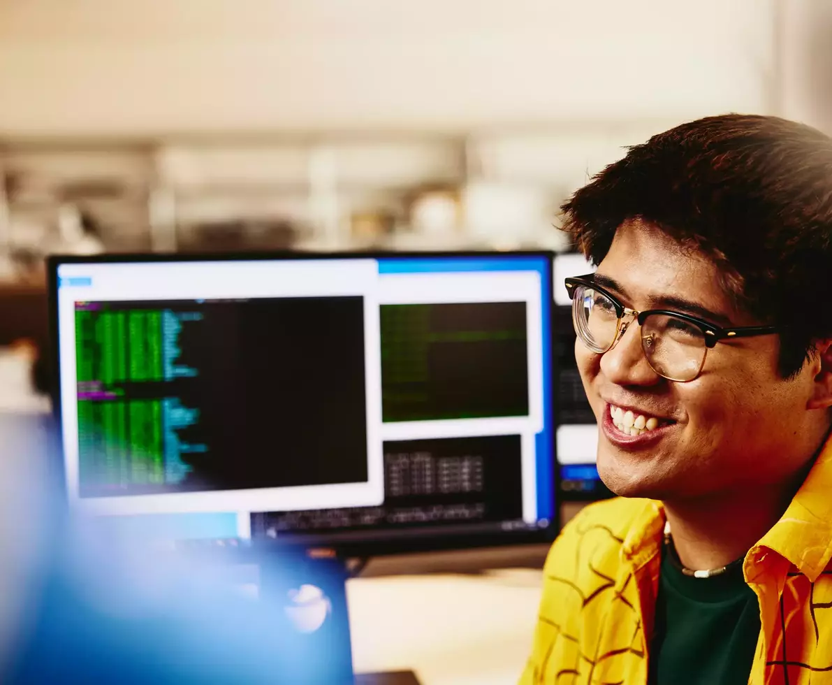 male smiling while sitting behind his desk, computer screens displaying programming code.
