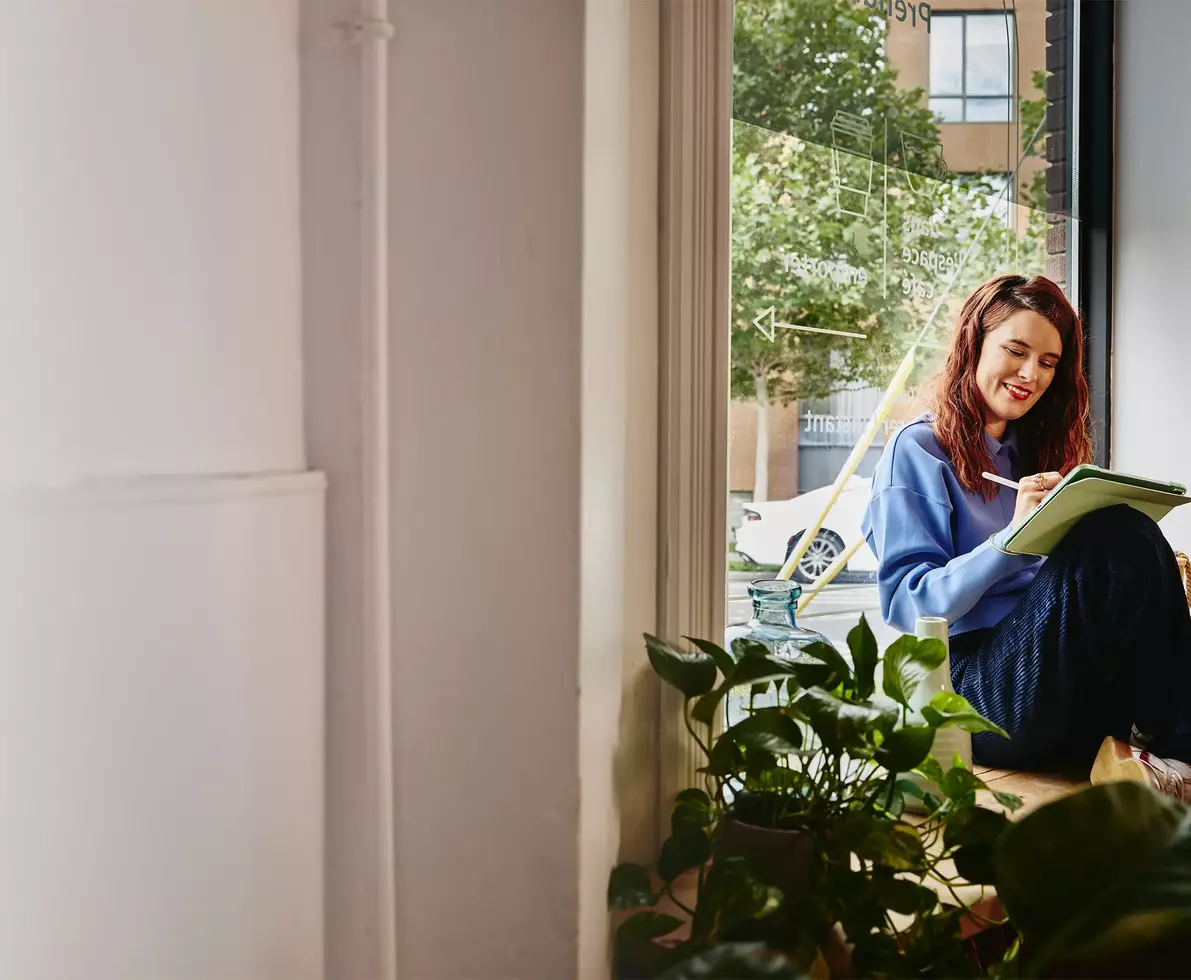 smiling female sitting next to a window, writing 